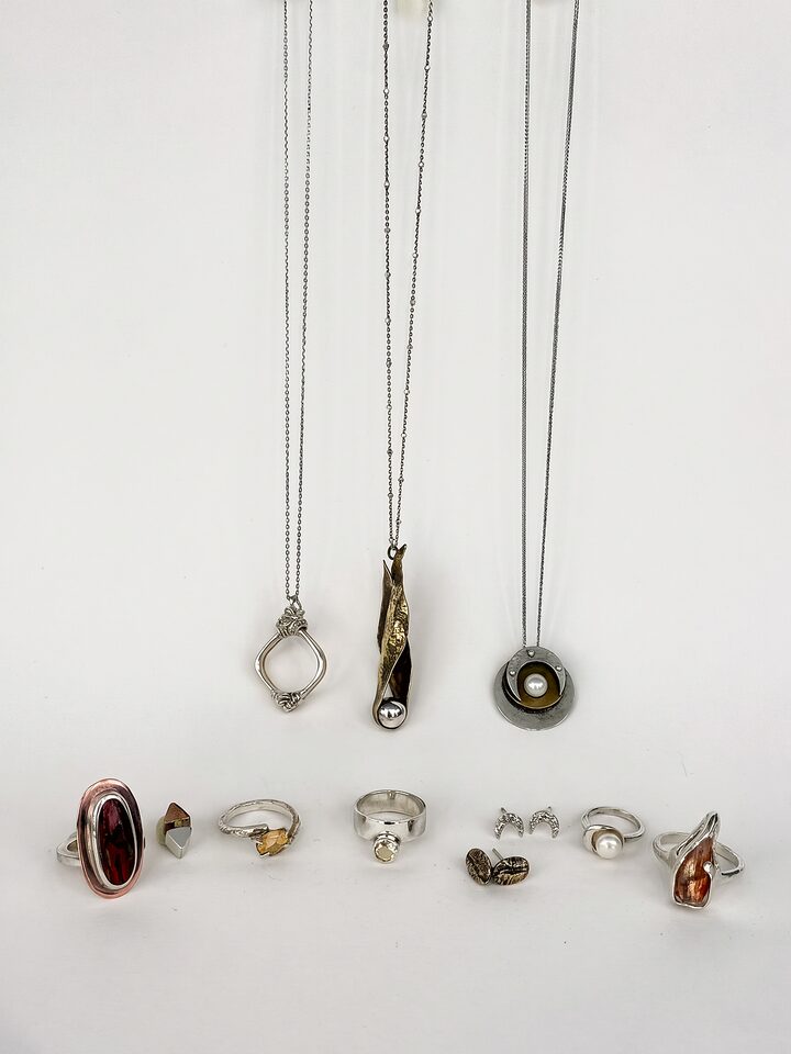 Silver and brass jewelry on white: three pendants on fine chains above rings with citrine, sunstone, pearl and a red stone, plus crescent and coffee bean studs.