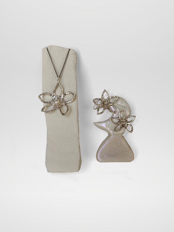 Silver flower pendant with a white pearl on a white display, beside two silver flower stud earrings.