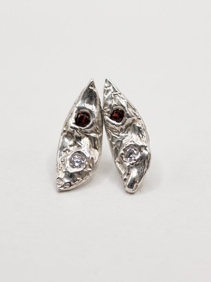 Pair of textured silver leaf stud earrings, each with a small red stone and a clear stone, on a white background.