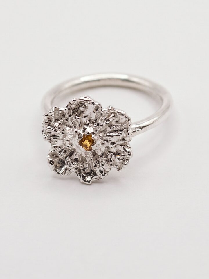 Silver flower ring with textured ruffled petals and a small yellow sapphire at the centre, on a round silver band, on white.