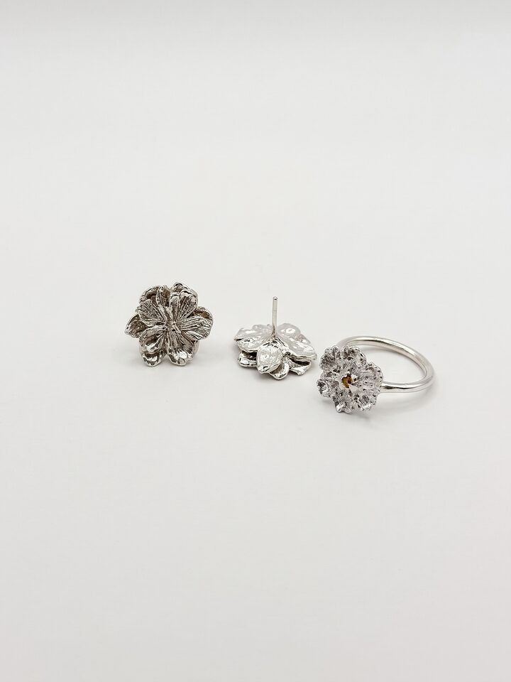 Two textured silver flower stud earrings, one turned to show its post, beside a silver flower ring with a small yellow sapphire, on white.