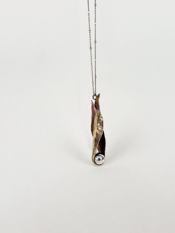 Twisted brass leaf pendant curled around a polished silver ball, hanging on a fine beaded silver chain, on white.