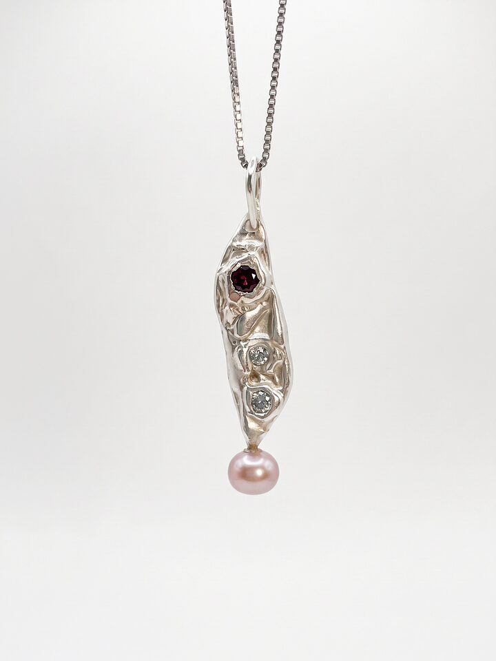 Long textured silver pendant with a small red stone and two small diamonds above a pink pearl drop, on a silver box chain, on white.