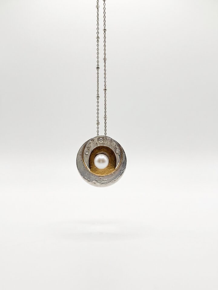 Round textured silver disc pendant with a white pearl on a golden brass disc, framed by a silver crescent with three tiny clear stones, on a beaded chain.