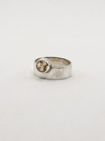 Wide flat silver band ring with a round pale yellow citrine raised at its edge, on a white background.
