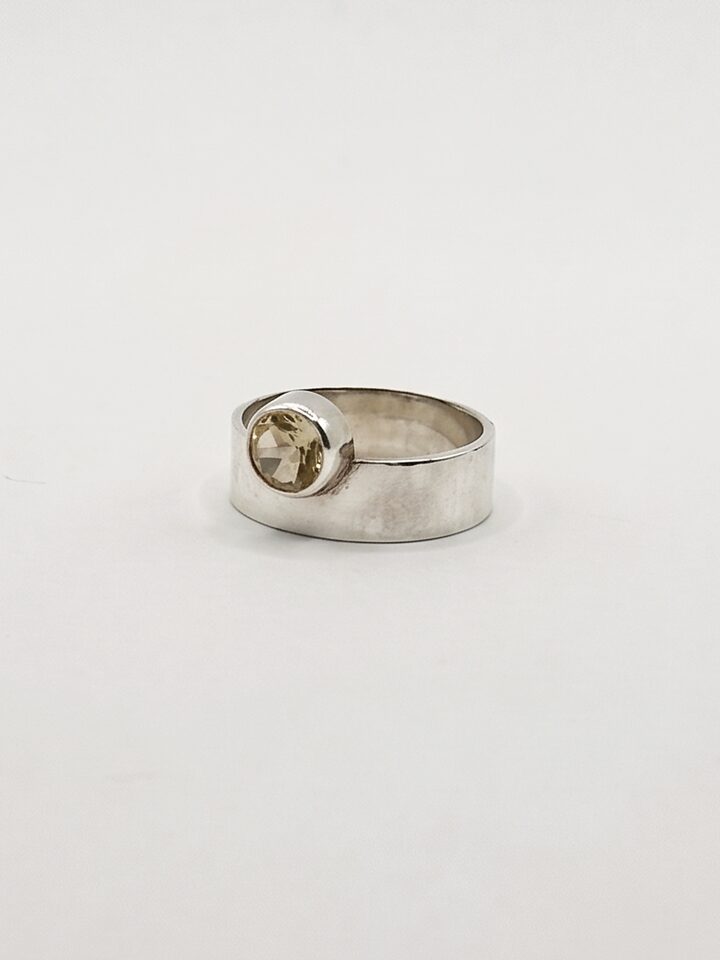 Wide flat silver band ring with a round pale yellow citrine raised at its edge, on a white background.