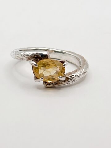 Silver bypass ring with an oval faceted citrine held by four slim claws on a textured band, on a white background.