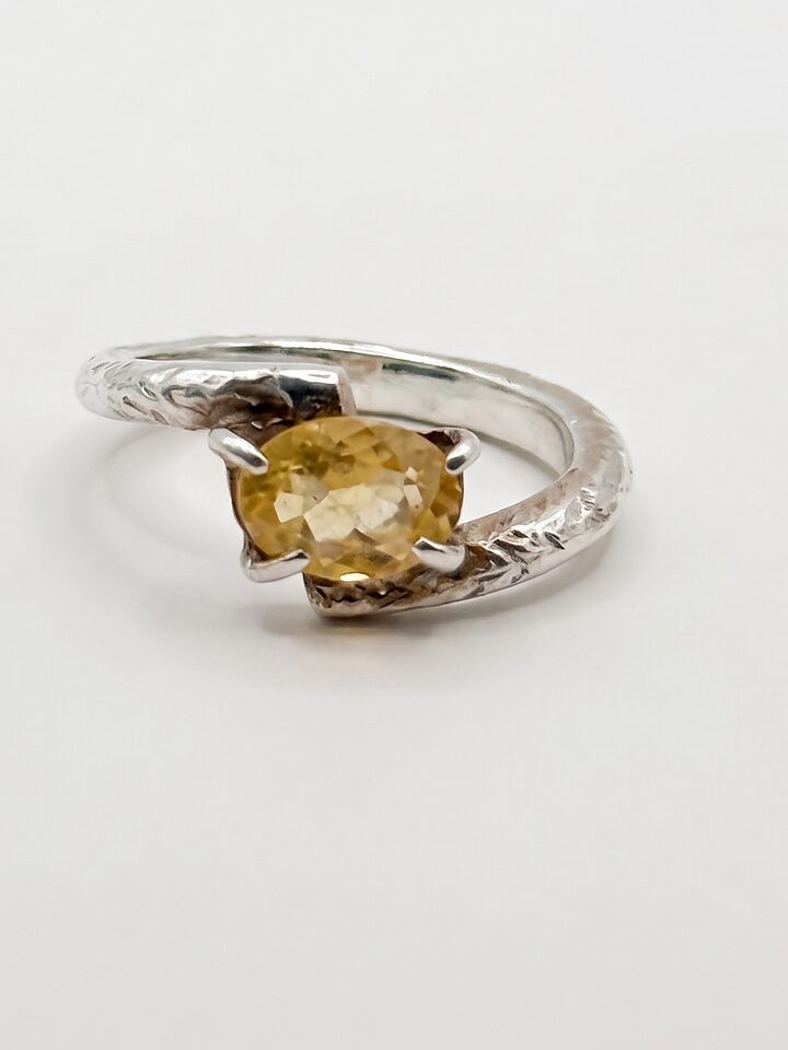 Silver bypass ring with an oval faceted citrine held by four slim claws on a textured band, on a white background.