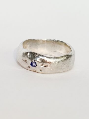 Wide organic silver band ring with a small blue stone inside engraved star rays, on a white background.