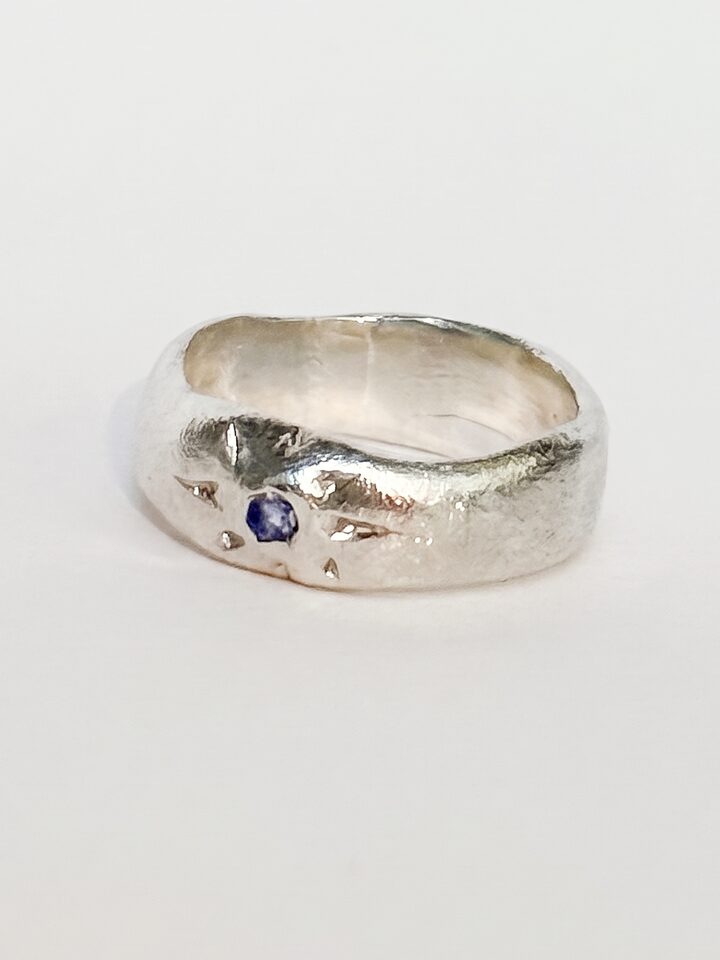 Wide organic silver band ring with a small blue stone inside engraved star rays, on a white background.
