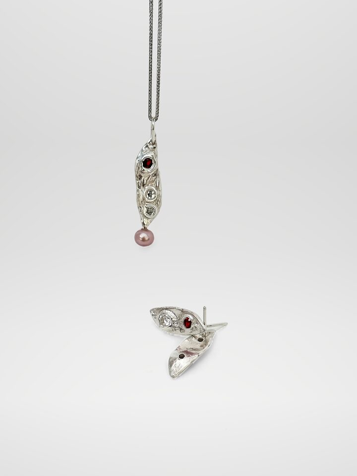 Long textured silver pendant with a red stone and two small diamonds above a pink pearl drop, with matching leaf studs holding a clear and a red stone, on white.