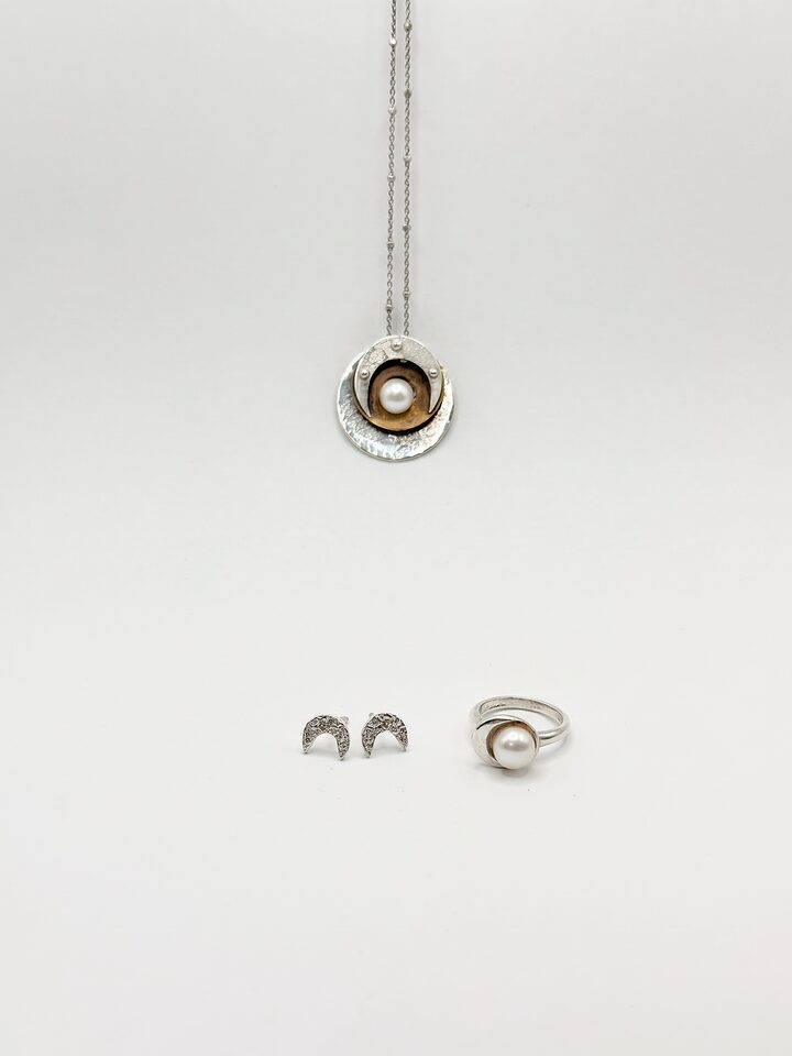 Silver disc pendant with a white pearl on a golden brass inner disc, crescent moon stud earrings and a silver pearl ring, on white.