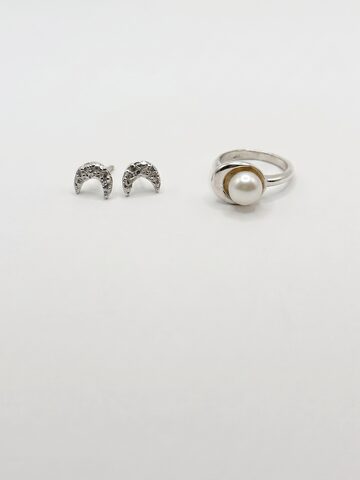 Textured silver crescent moon stud earrings beside a silver ring holding a white pearl in a crescent cup, on white.