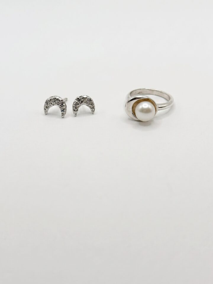 Textured silver crescent moon stud earrings beside a silver ring holding a white pearl in a crescent cup, on white.